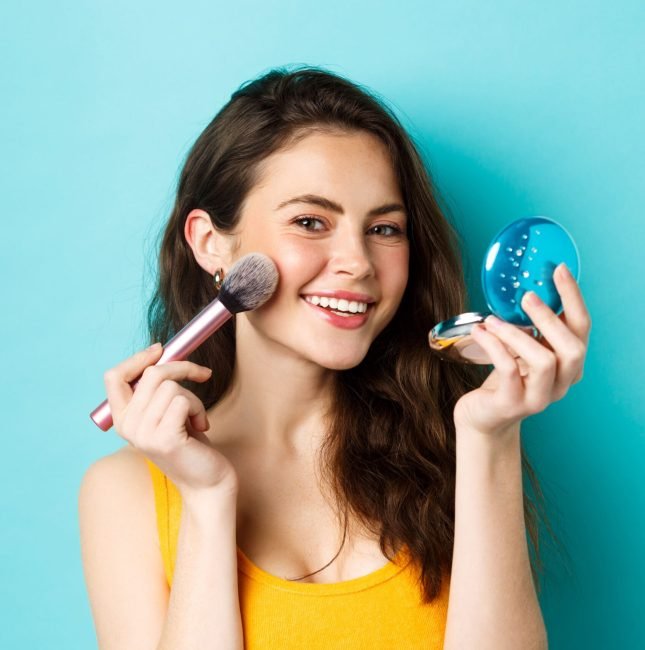 beauty-close-up-attractive-woman-looking-mirror-using-brush-apply-make-up-smiling-pleased-camera-standing-against-blue-background